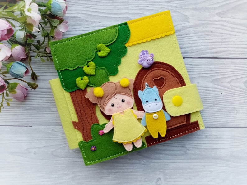 May include: A felt book with a green tree, a yellow house, a girl with brown hair wearing a yellow dress, and a blue unicorn wearing a yellow shirt. The book has a brown door with a heart on it and a yellow button.