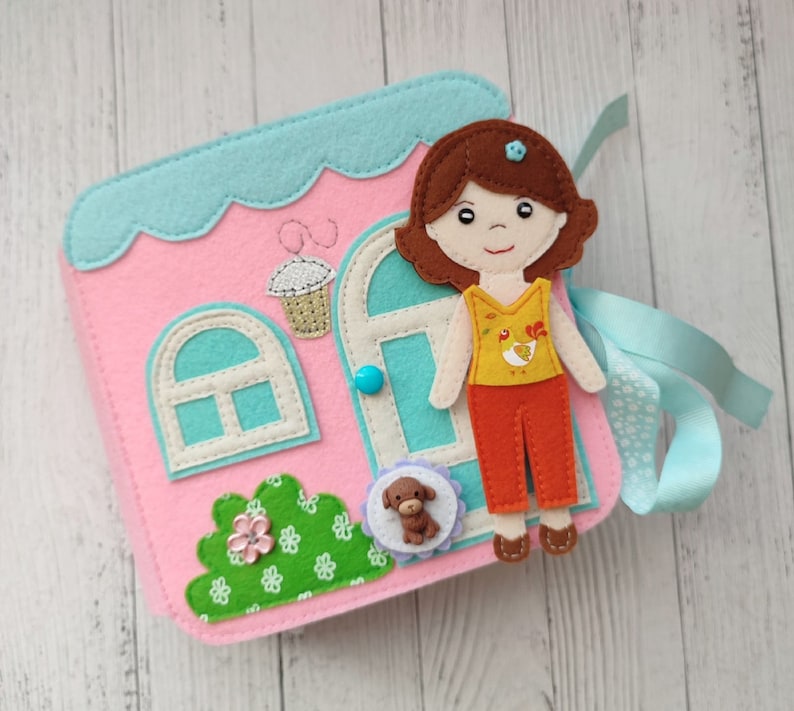 May include: A felt dollhouse playset with a girl doll, a dog, and a window with a blue button. The dollhouse is pink with a blue roof and a green bush.