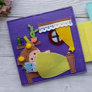 May include: A felt book page with a purple background featuring a girl in a brown hat and yellow pajamas sleeping in a bed with a green blanket. The girl is holding a blue stuffed animal. There is a window with a yellow curtain and a bookshelf with books on the wall.
