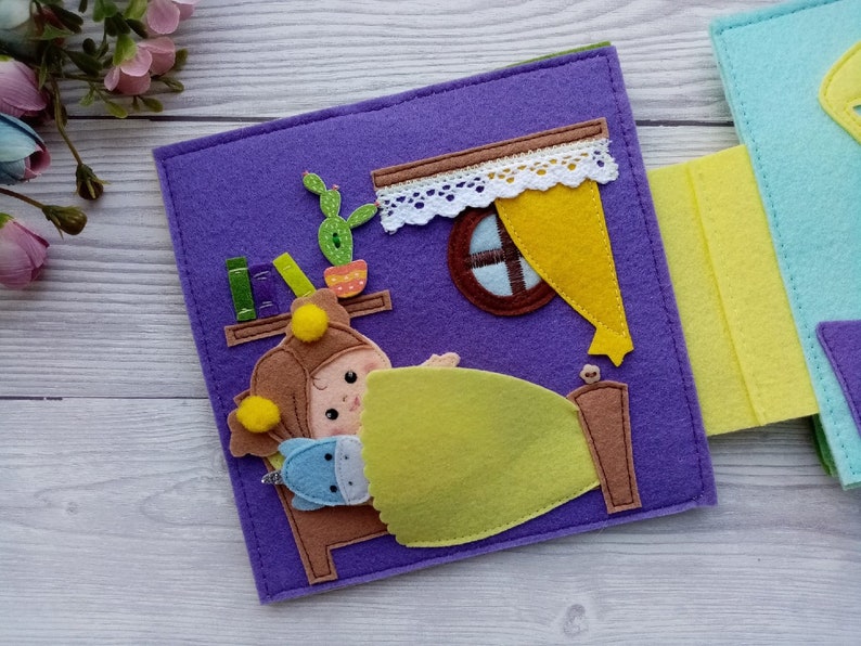 May include: A felt book page with a purple background featuring a girl in a brown hat and yellow pajamas sleeping in a bed with a green blanket. The girl is holding a blue stuffed animal. There is a window with a yellow curtain and a bookshelf with books on the wall.