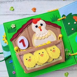 Pode incluir: Uma p&aacute;gina de livro de feltro com fundo verde, um galinheiro marrom com telhado vermelho, uma galinha branca com crista vermelha e um pintinho amarelo dentro de uma bolsa de malha. O n&uacute;mero "1" est&aacute; no galinheiro.