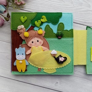 May include: A felt book with a girl, a blue and yellow animal, and a teapot with a cupcake on a table. The book is open to a page with a green background and a forest scene.