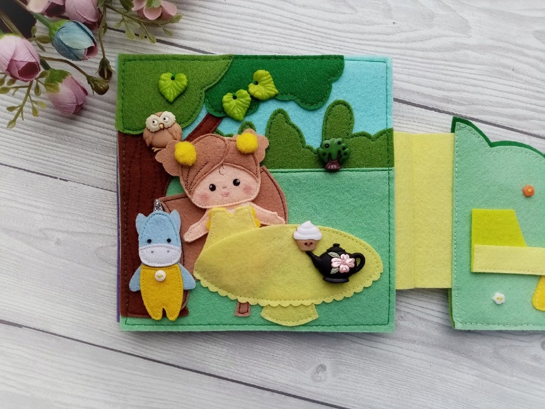 May include: A felt book with a girl, a blue and yellow animal, and a teapot with a cupcake on a table. The book is open to a page with a green background and a forest scene.