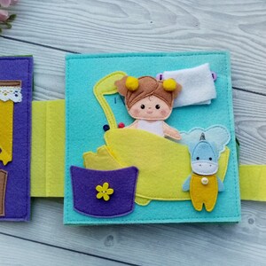 May include: A felt book page with a girl doll in a yellow bathtub, a unicorn toy, and a purple bucket with a yellow flower. The page is light blue with yellow felt trim.