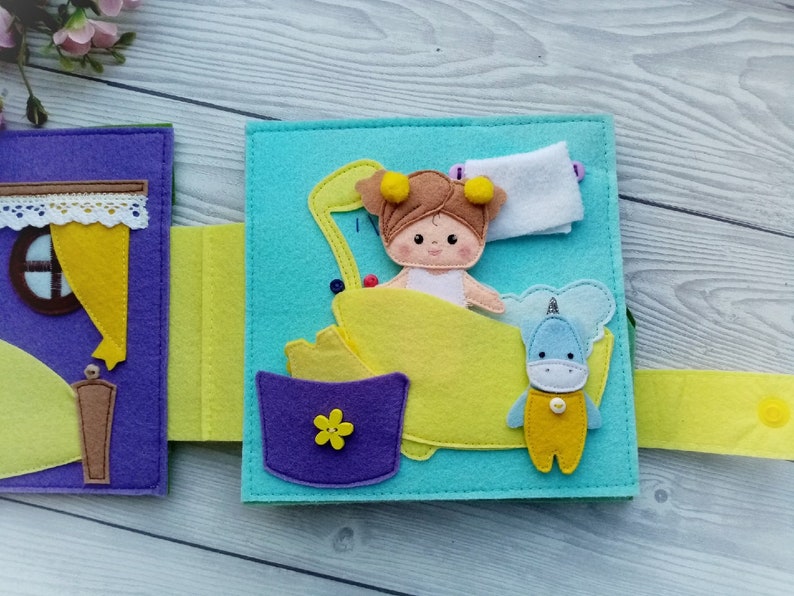 May include: A felt book page with a girl doll in a yellow bathtub, a unicorn toy, and a purple bucket with a yellow flower. The page is light blue with yellow felt trim.