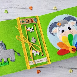 Pode incluir: Um livro de feltro verde com uma borda amarela e um animal de feltro branco em cada p&aacute;gina. O livro tem uma atividade de amarra&ccedil;&atilde;o com um cord&atilde;o amarelo e uma variedade de formas de feltro. O texto "Handmade" &eacute; vis&iacute;vel no livro.