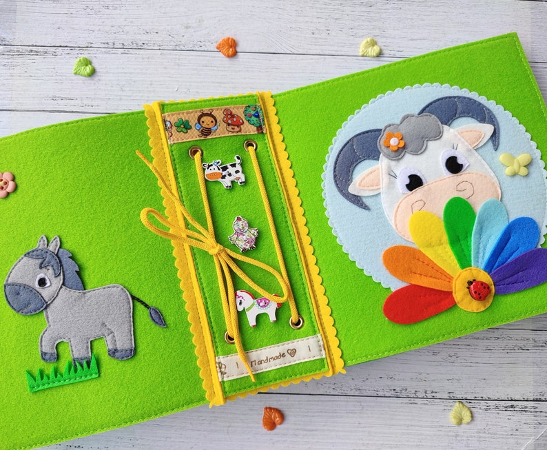 Pode incluir: Um livro de feltro verde com uma borda amarela e um animal de feltro branco em cada p&aacute;gina. O livro tem uma atividade de amarra&ccedil;&atilde;o com um cord&atilde;o amarelo e uma variedade de formas de feltro. O texto "Handmade" &eacute; vis&iacute;vel no livro.