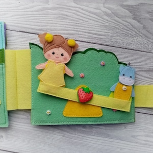 May include: A felt book with a green, yellow, and blue color scheme. The book features a girl with yellow hair and a yellow dress, a blue unicorn, and a strawberry. The book is designed for children.