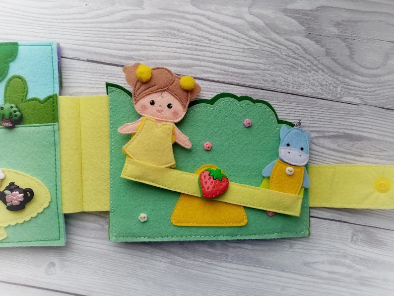 May include: A felt book with a green, yellow, and blue color scheme. The book features a girl with yellow hair and a yellow dress, a blue unicorn, and a strawberry. The book is designed for children.