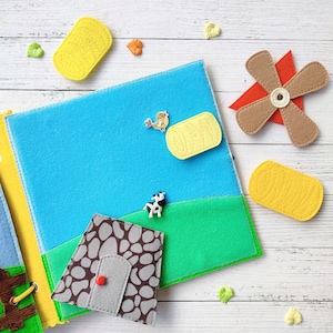 Pode incluir: Um livro de feltro com um c&eacute;u azul, grama verde, uma casa marrom, uma fardo de feno amarelo, um moinho de vento marrom e uma vaca branca. O livro est&aacute; aberto em uma p&aacute;gina com um c&eacute;u azul e um campo verde.