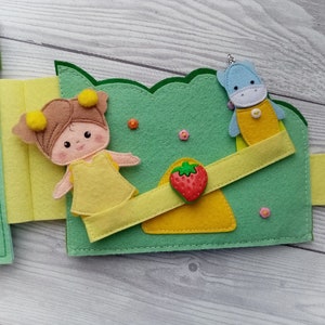 May include: A felt book page with a girl doll, a unicorn doll, and a strawberry on a seesaw. The dolls are wearing yellow dresses and the seesaw is yellow and green.
