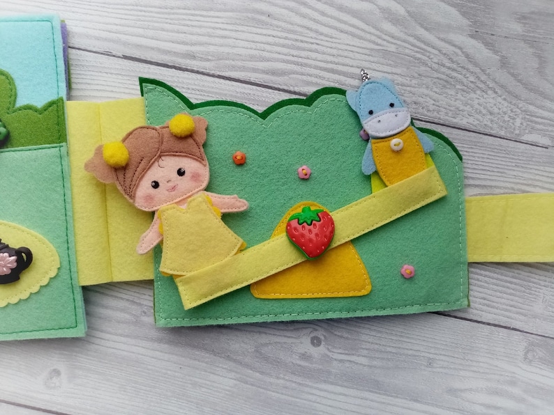 May include: A felt book page with a girl doll, a unicorn doll, and a strawberry on a seesaw. The dolls are wearing yellow dresses and the seesaw is yellow and green.