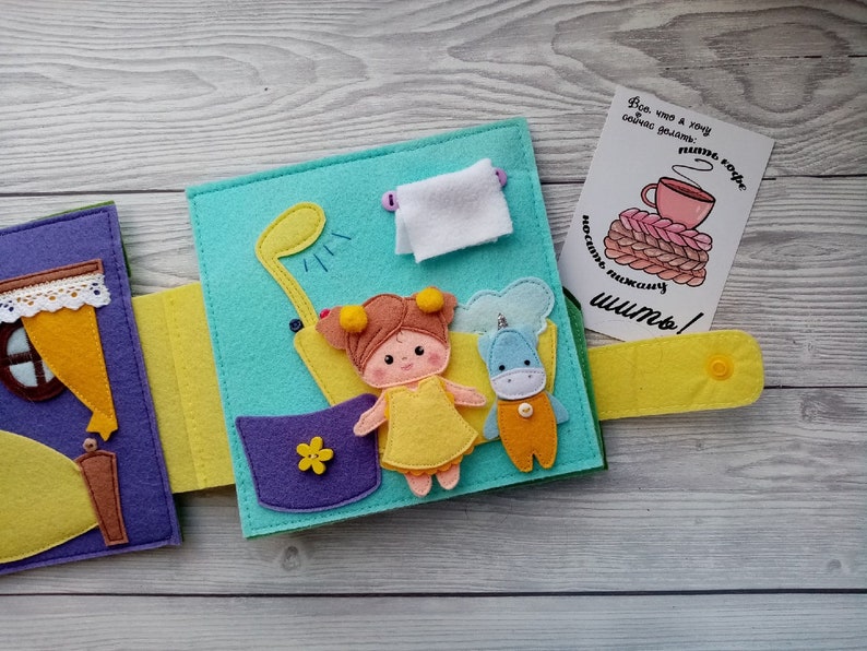May include: A felt book for children with a purple, yellow, and blue cover. The book has a variety of felt characters and objects, including a girl, a unicorn, a shower, and a cup of coffee. The text on the book says "What I want to do now: sew!"