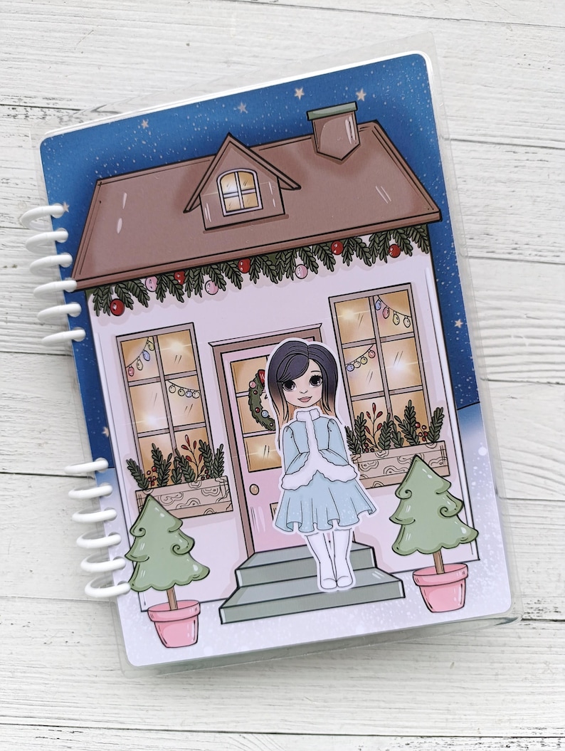 Pu&ograve; includere: Una casa bianca con un tetto marrone e un cielo blu con stelle. La casa ha una porta rosa e due finestre con luci gialle. Ci sono due alberi di Natale verdi in vaso davanti alla casa. Una ragazza con lunghi capelli neri &egrave; in piedi sui gradini davanti alla casa. Indossa un vestito blu con un bordo di pelliccia bianca e calze bianche.