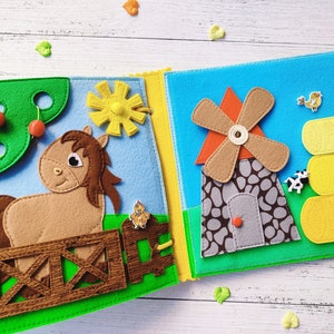 Pode incluir: Um livro de feltro para crian&ccedil;as com duas p&aacute;ginas. A p&aacute;gina da esquerda apresenta um cavalo castanho em p&eacute; atr&aacute;s de uma cerca de madeira, uma &aacute;rvore verde com cerejas vermelhas e um sol amarelo. A p&aacute;gina da direita apresenta um moinho de vento cinza com uma l&acirc;mina laranja, uma fardo de feno amarelo e uma pequena vaca branca.