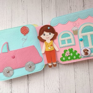 May include: A felt book for children with a pink car, a girl doll, and a house with a blue door. The book has a light blue background and is decorated with colorful details.