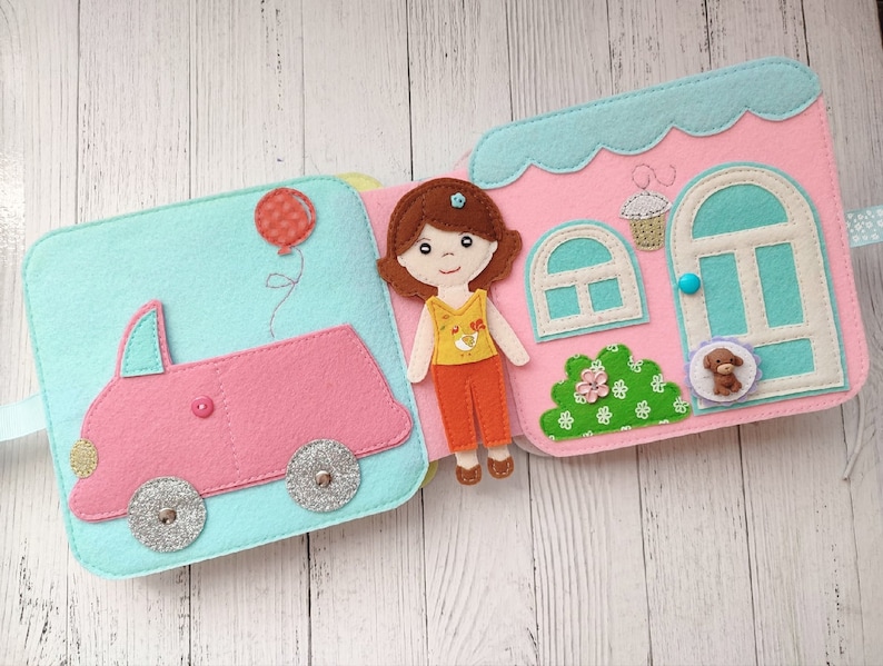 May include: A felt book for children with a pink car, a girl doll, and a house with a blue door. The book has a light blue background and is decorated with colorful details.