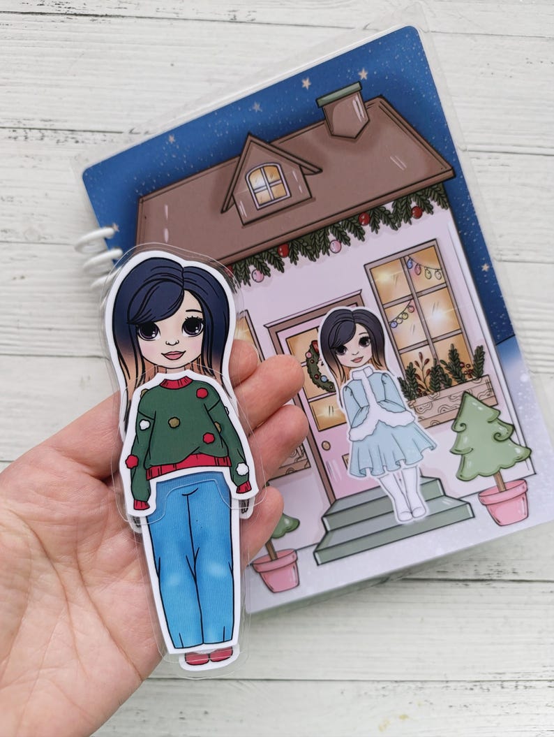 Pu&ograve; includere: Un set di due bambole di carta a tema invernale. Le bambole sono vestite con abiti festivi. Lo sfondo &egrave; una casa con una porta rosa e un albero di Natale.