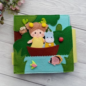 May include: A felt book for children featuring a girl and a small animal in a boat on a lake. The book has a green, blue, and yellow color scheme and includes a ladybug, an owl, and fish.