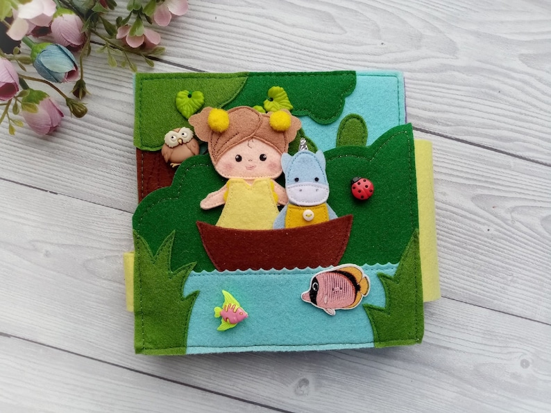 May include: A felt book for children featuring a girl and a small animal in a boat on a lake. The book has a green, blue, and yellow color scheme and includes a ladybug, an owl, and fish.