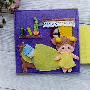 May include: A felt book page with a purple background featuring a girl doll in a yellow dress standing next to a bed with a unicorn doll tucked in. The bed has a brown headboard and a yellow blanket. There is a cactus and a bookshelf with books on the wall behind the bed.