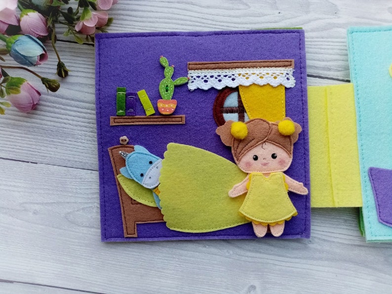 May include: A felt book page with a purple background featuring a girl doll in a yellow dress standing next to a bed with a unicorn doll tucked in. The bed has a brown headboard and a yellow blanket. There is a cactus and a bookshelf with books on the wall behind the bed.