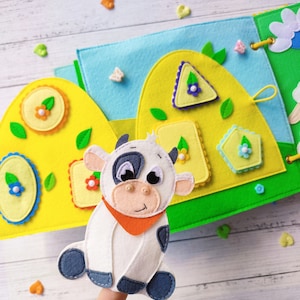Pode incluir: Um fantoche de dedo de feltro de uma vaca branca com manchas pretas e uma bandana laranja. O fantoche est&aacute; sentado em um dedo e est&aacute; na frente de um livro de feltro com formas e flores coloridas.