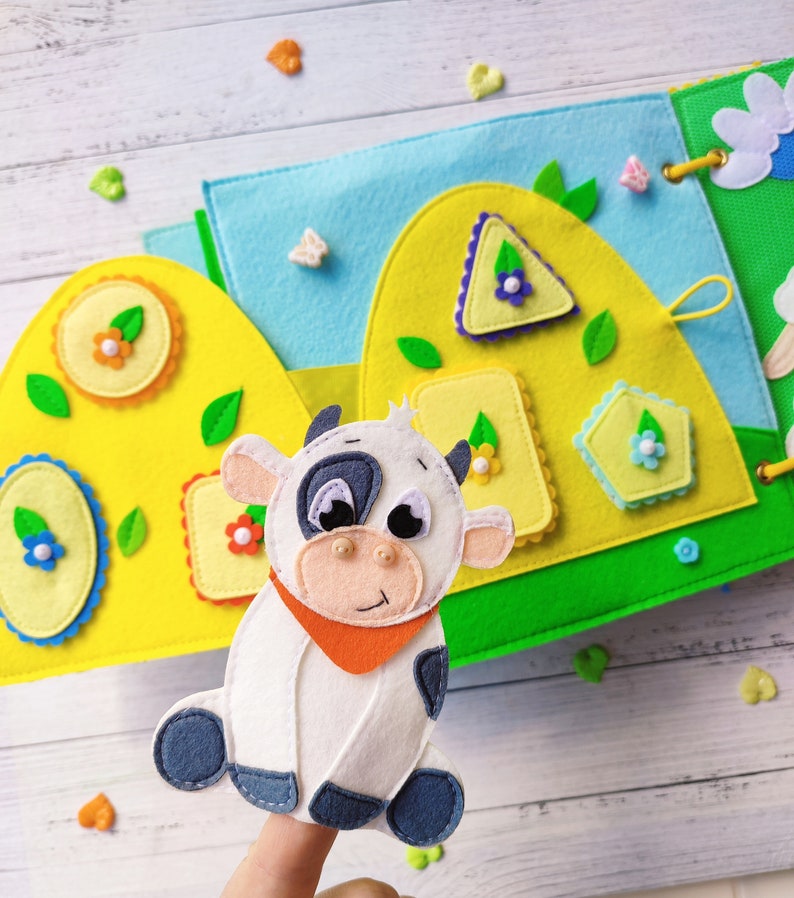 Pode incluir: Um fantoche de dedo de feltro de uma vaca branca com manchas pretas e uma bandana laranja. O fantoche est&aacute; sentado em um dedo e est&aacute; na frente de um livro de feltro com formas e flores coloridas.