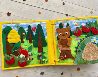 May include: A felt book for children with a forest theme. The book features a beehive, trees, a hedgehog, a bear, and a mushroom. The book is yellow with white polka dots.