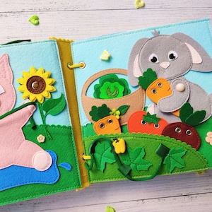 Pode incluir: Um livro de feltro para crian&ccedil;as com um porco rosa em uma p&aacute;gina e um coelho cinza na outra. O porco est&aacute; segurando uma corda verde e o coelho est&aacute; segurando uma cenoura. As p&aacute;ginas s&atilde;o decoradas com flores, vegetais e folhas de feltro.