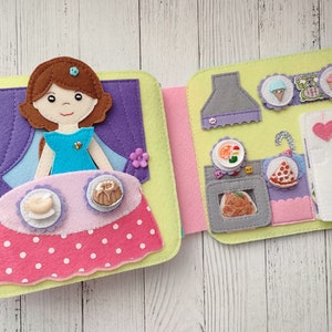 May include: A felt book for children with a girl doll on the front cover and a kitchen scene on the inside. The kitchen scene includes a stove, oven, refrigerator, and various food items.