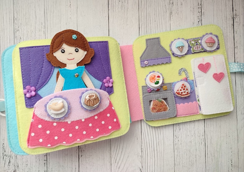 May include: A felt book for children with a girl doll on the front cover and a kitchen scene on the inside. The kitchen scene includes a stove, oven, refrigerator, and various food items.