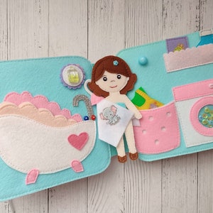 May include: A felt play set for children featuring a doll, a bathtub, a laundry basket, and a washing machine. The doll is holding a towel with an elephant design. The bathtub is pink and white with a heart on the side. The laundry basket is pink with a green shirt inside. The washing machine is white with a pink lid and a clear window showing colorful sequins.