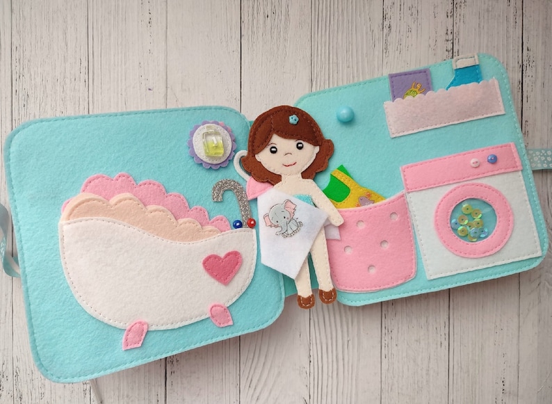May include: A felt play set for children featuring a doll, a bathtub, a laundry basket, and a washing machine. The doll is holding a towel with an elephant design. The bathtub is pink and white with a heart on the side. The laundry basket is pink with a green shirt inside. The washing machine is white with a pink lid and a clear window showing colorful sequins.