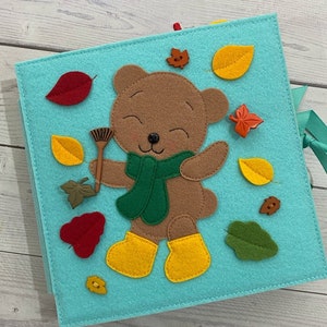 May include: A felt book featuring a brown bear wearing a green scarf and yellow boots. The bear is holding a rake and there are leaves and buttons on the cover.