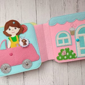May include: A felt book with a pink car and a girl holding a teddy bear on the left page. The right page shows a pink house with a blue door and a window.