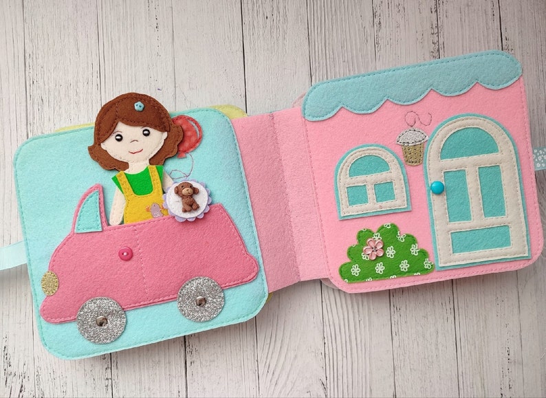 May include: A felt book with a pink car and a girl holding a teddy bear on the left page. The right page shows a pink house with a blue door and a window.