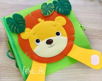 Op de afbeelding: Een viltboek voor kinderen met een leeuw met een geel gezicht, een oranje manen en gele poten. De leeuw is omringd door groene viltbladeren. Het boek heeft de tekst "Fetro_fei" op de voorkant.