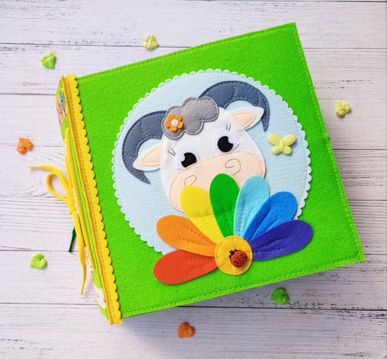 Pode incluir: Um livro de feltro verde com um animal de feltro branco e uma flor na capa. O animal tem um rosto branco com olhos pretos e um nariz preto. A flor &eacute; arco-&iacute;ris com uma joaninha vermelha no centro.