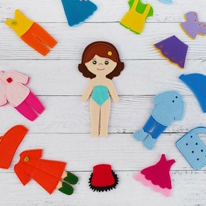 May include: Felt doll with brown hair and a blue dress, surrounded by various felt clothing items including a pink shirt, a yellow dress, a blue hat, and a red coat.