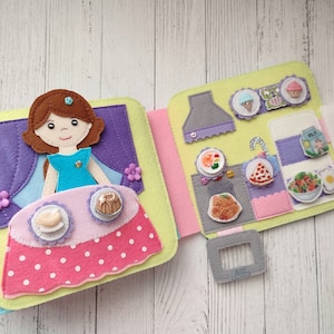 May include: A felt book with a doll and food. The doll is wearing a blue dress and has brown hair. The food includes a pizza, pasta, salad, and ice cream. The book is open to show the inside pages.