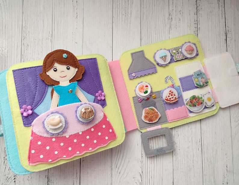 May include: A felt book with a doll and food. The doll is wearing a blue dress and has brown hair. The food includes a pizza, pasta, salad, and ice cream. The book is open to show the inside pages.