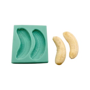 May include: A light green silicone banana mold with two banana-shaped cavities, next to two peeled bananas. The mold is square and the bananas are a light yellow color.