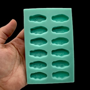 May include: A teal silicone mold with twelve car-shaped cavities. Each cavity has a detailed car design. The mold is held in a hand against a black background. The car molds are for crafting.