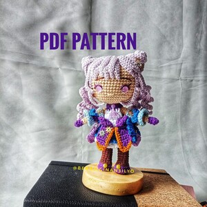 Puede incluir: Una muñeca amigurumi de crochet con cabello morado, un vestido azul y morado y una estrella amarilla en el pecho. La muñeca está de pie sobre una base de madera y el texto "PDF PATTERN" está escrito en letras moradas sobre la muñeca.
