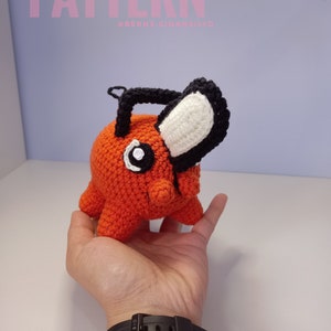 Könnte beinhalten: Ein gehäkeltes Plüschtier in Orange und Schwarz mit weißen Akzenten. Das Spielzeug hat einen großen Kopf mit einem schwarzen Ohr und einem weißen Ohr. Das Spielzeug wird in der Hand einer Person gehalten.