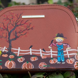 May include: A brown leather crossbody bag with a painted design of a scarecrow in a field of pumpkins. The scarecrow is wearing a blue jumpsuit and a straw hat. There is a tree with red leaves in the background.