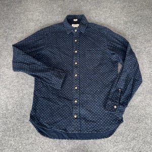 May include: A long-sleeve button-down shirt in dark navy blue featuring a repeating pattern of small white dots. The shirt includes a front chest pocket, a standard collar, and light-colored buttons running down the center placket and on the cuffs.