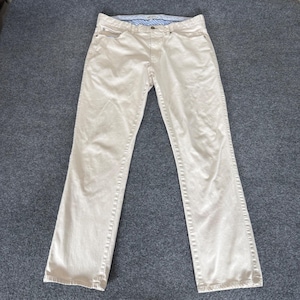 May include: A pair of light tan-colored denim jeans. The jeans have a classic five-pocket design, a button and zipper closure, and a straight leg fit. The jeans are laid flat on a gray surface.
