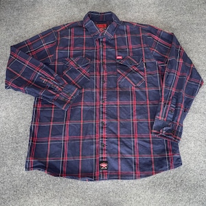 May include: A long-sleeved, button-down shirt in a plaid pattern of navy blue, red, and white. The shirt has two chest pockets and a small logo patch near the bottom. The brand name "Dixon" is visible on the collar.